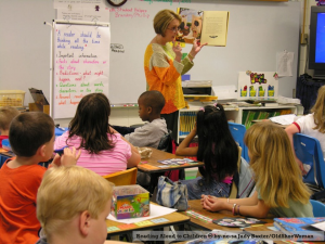 Photo of teacher in a classroom holding a book up as she stands before her 4th-or-5th-grade-sized students at their desks