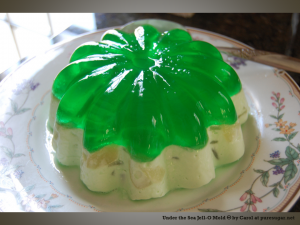 Jello mold on a plate, the bottom half is opaque and cream-colored, with embedded bits of other food; the top layer is clear green jello