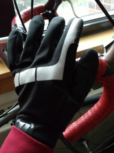 Full hand in lightweight black glove with reflective gray stripe. Hand is resting on bike handlebars, which are leaning against a windowsill in the background.