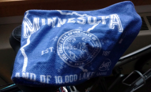 Light blue square of fabric draped over barely visible narrow black bike seat. The fabric has white printing with an outline of the state of Minnesota, and the state seal in the center.