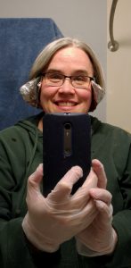 White woman with pale pink skin, light gray straight hair, and glasses) standing, pointing phone camera at a bathroom mirror. She (me) is smiling, and wearing cheezy plastic gloves, and has a piece of tinfoil wrapped behind the back of her neck, and over her ears. The tinfoil curves away from her face like very dorky wings just in front of her ears.