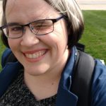 White woman with pale pink skin and chin-length grey hair with black tips standing outdoors. She's wearing a patterned black shirt, a blue blazer, and a backpack. She's squinting into the sun with her left eye. Behind her you can see grass, and part of Notre Dame's stadium.