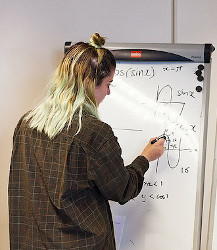 White woman with long multi-hued hair faces a whiteboard, working out a math problem