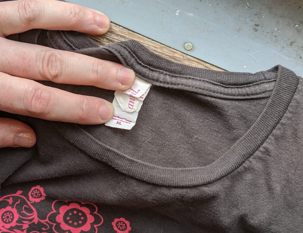 Close view of the collar of a grey t-shirt. A hand is holding down the tag to show the brand name "Anvil" and size XL