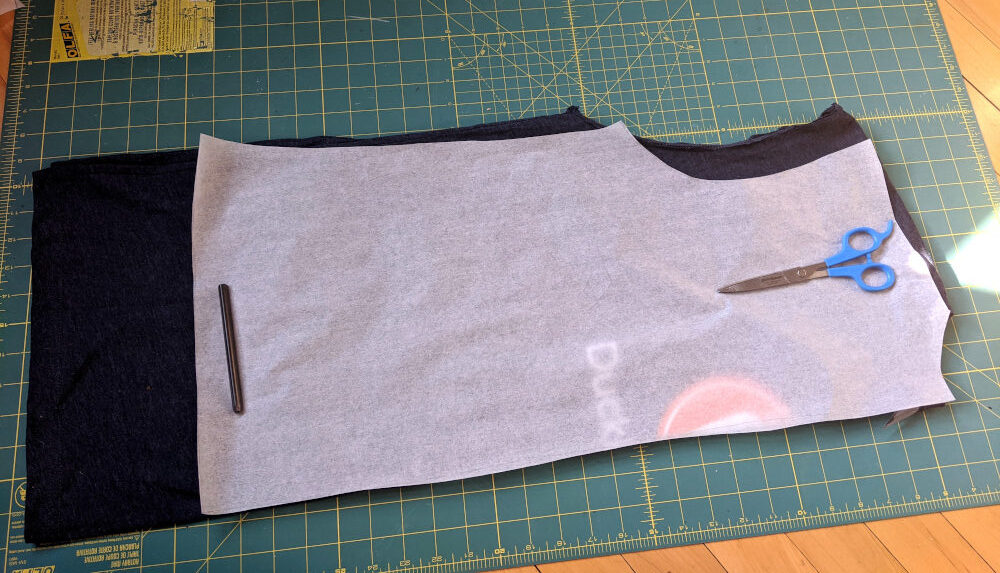 A dark navy t-shirt with its sleeves cut off, has been folded in half with the front of the shirt facing out. A sewing pattern piece for the front of a t-shirt is lying on top of the shirt - it's one of the pieces obtained by tracing in the previous step.