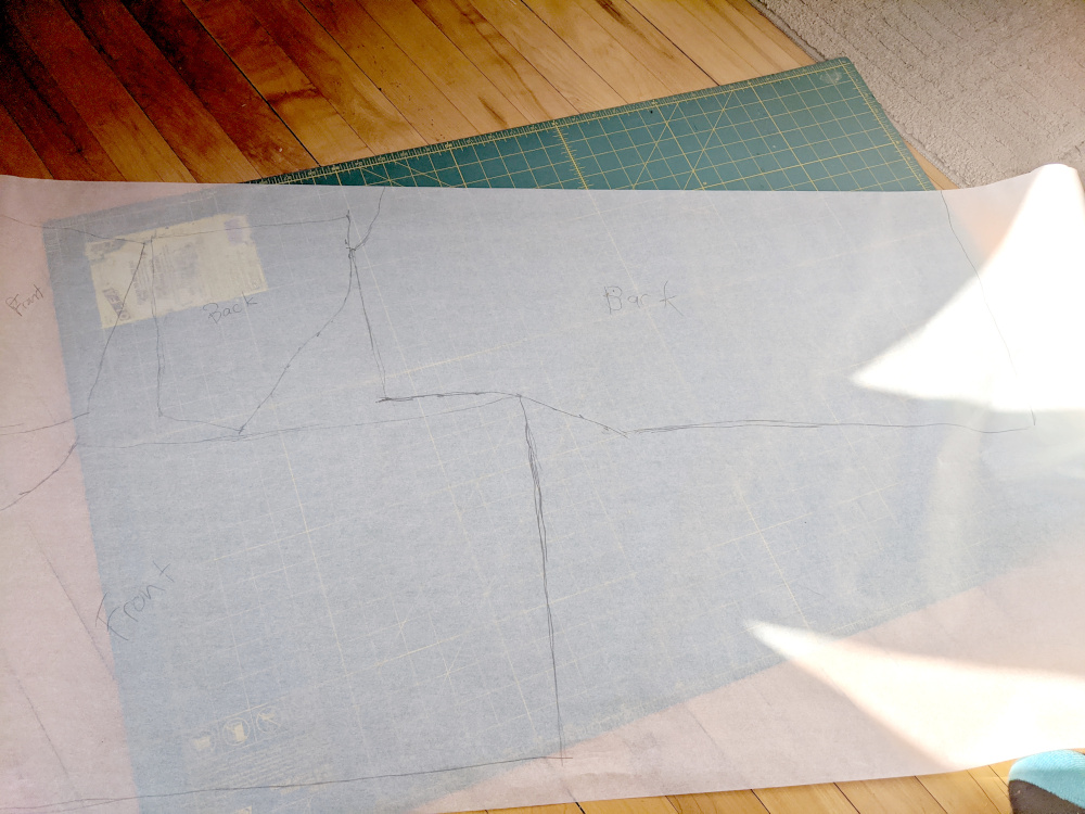 A large piece of tracing paper lying on the floor, on a green cutting mat. The outlines of the front and back halves of a t-shirt, and the front and back of the sleeves, have been traced onto the paper.
