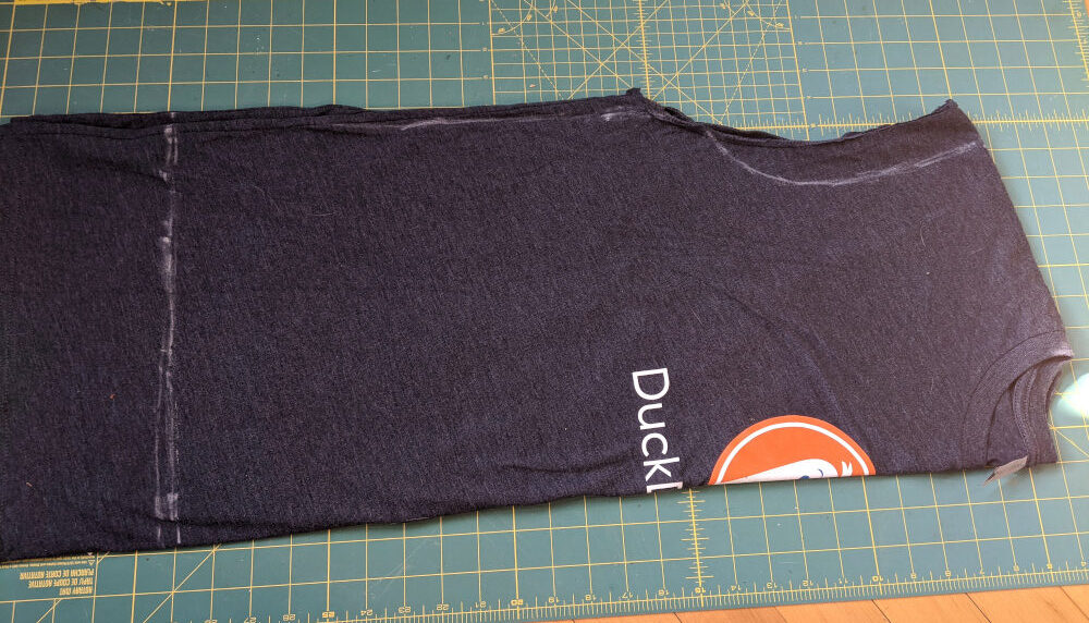 Dark navy t-shirt with the sleeves cut off, folded in half and lying on the floor, with chalk lines drawn on it to outline the pattern piece from the previous step.