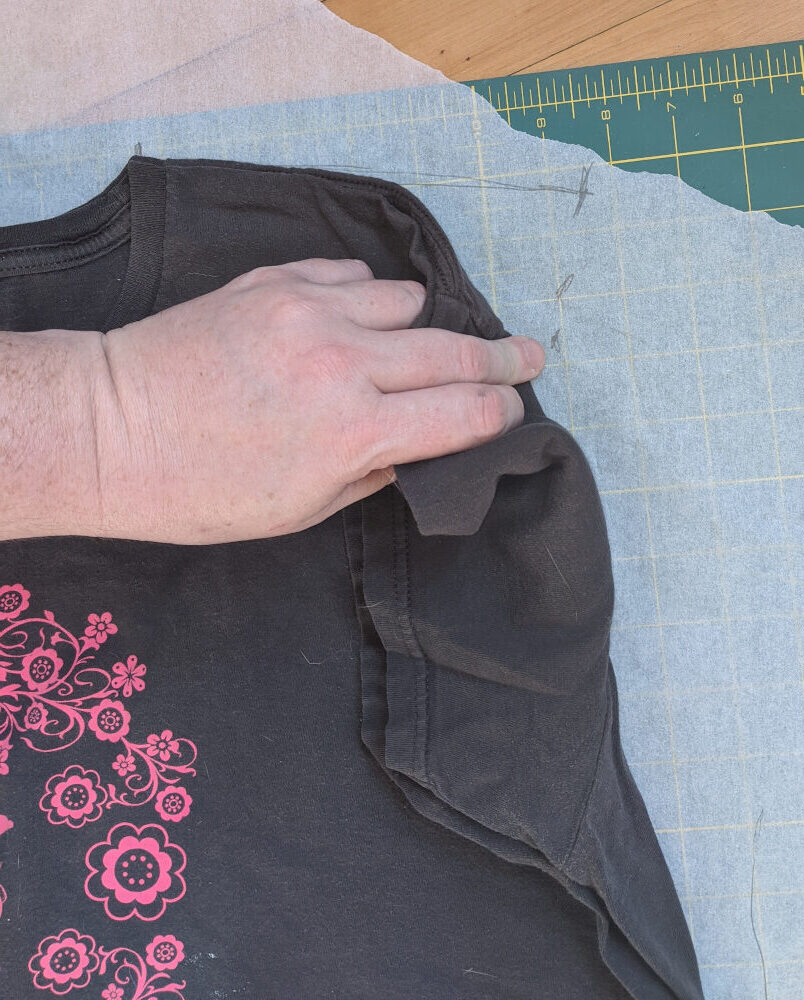 Grey t-shirt lying on a white piece of tracing paper (which is itself lying on a green cutting mat.) The top shoulder seam and side seam of the shirt have been traced on the paper, and a hand is pulling back the sleeve to show how to trace the edge of the arm hole.